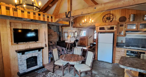 Cozy cabin interior featuring wooden beams, a dining table, fireplace, and modern kitchen appliances.